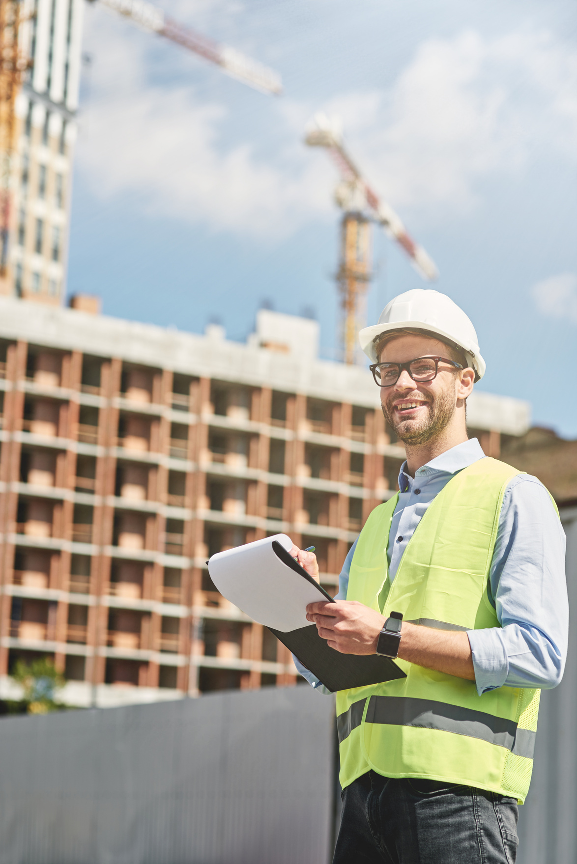 Good job. Young happy civil engineer or construction supervisor wearing helmet looking away and smiling while inspecting building site and making some notes